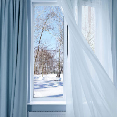 Light Blue And Sheer Window Curtains Installed On A Window, Snowing Outside. Insulating Blinds For Winter.
