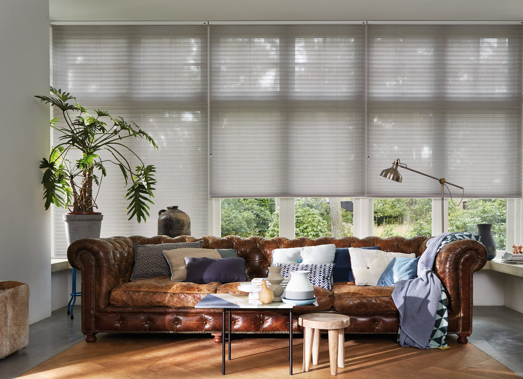 Eclectic style living room with wooden floors and large leather sofa, sheer Luxaflex Duette Shades partially covering large window panes, filtering through sun light. Made in Australia.