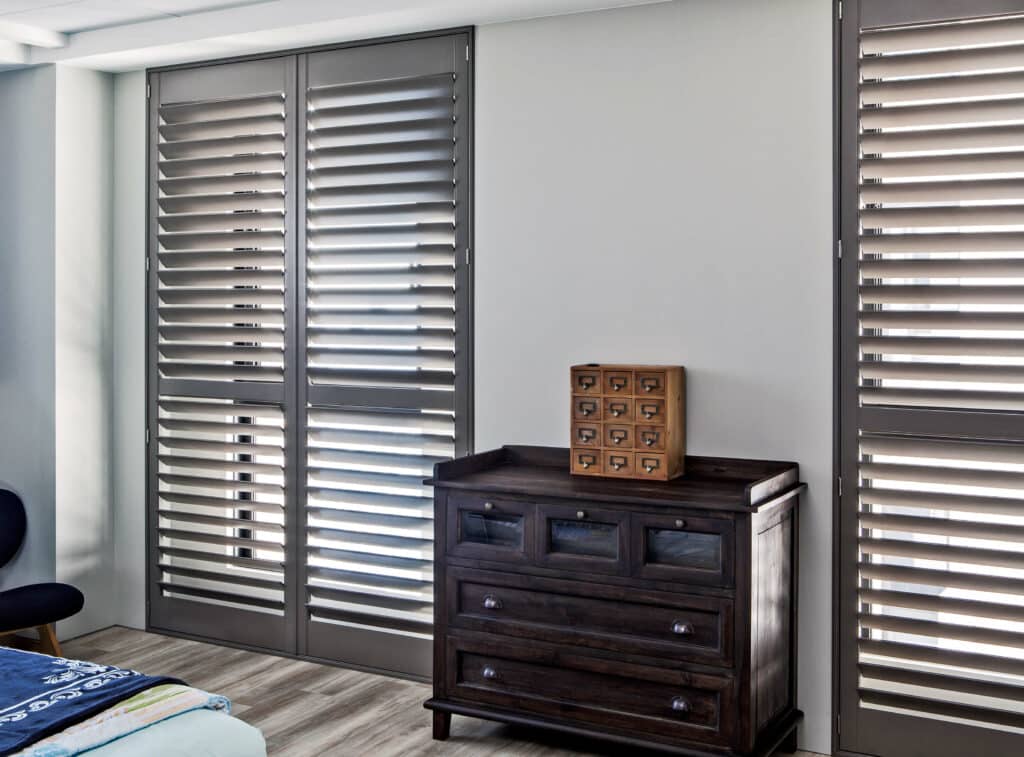 Dark grey Plantation Shutters in a bed room with an antique drawer. Modern and contemporary aesthetic. Shutters available at Complete Blinds Sydney. Plantation Shutter Colours.