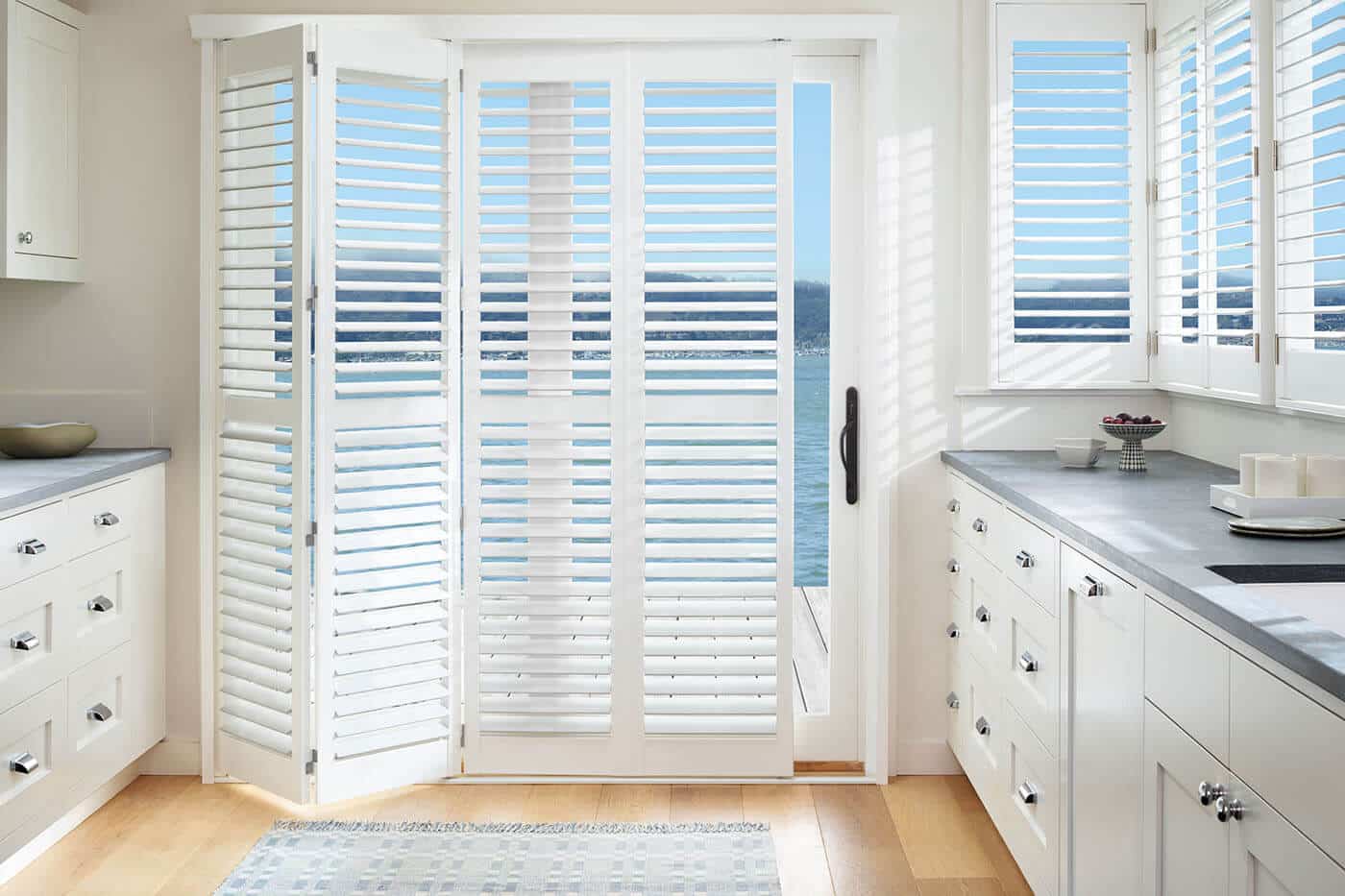 Transitional style kitchen with a glass sliding door revealing a patio and beach view, partially covered by Luxaflex Polysatin Shutters. Made in Australia. Water resistant.