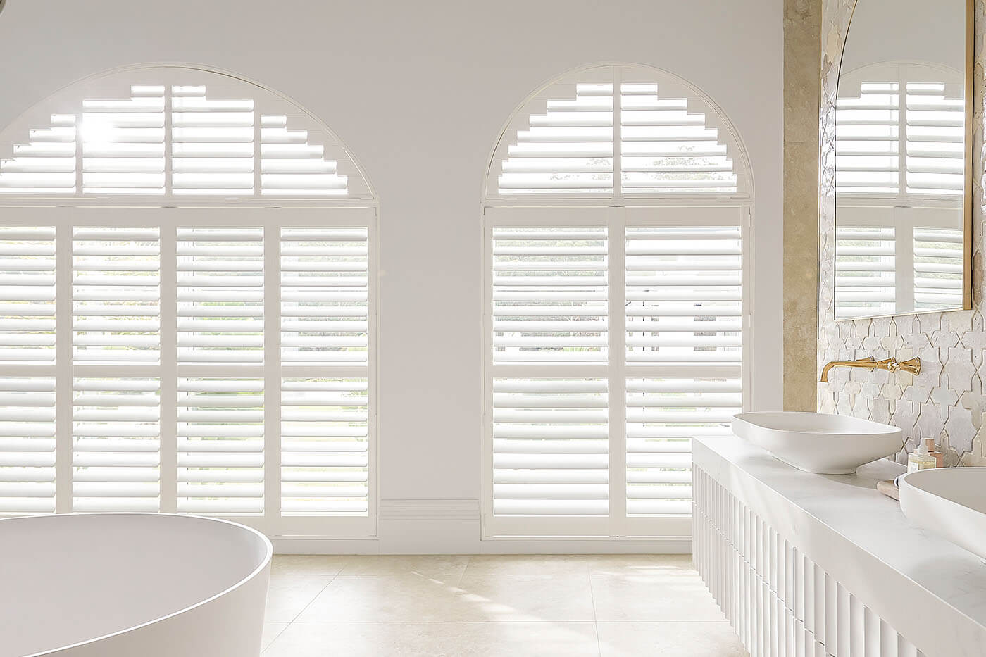 Classic transitional style white bathroom with large radius windows covered by Luxaflex Polysatin Shutters, offering light and privacy control in the bathroom. Suitable for wet areas. For sale in our Sydney showroom.