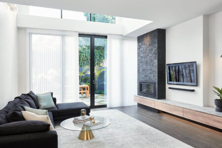 Minimal Living Room Design With White Walls, Black Chimney And Couch Featuring Luxaflex Luminette Privacy Sheers Covering A Large Window Showing The Outside House Garden.