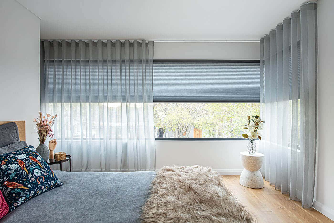 Modern transitional style blue-themed bedroom featuring long horizontal window panes partially covered by layers if Luxaflex curtain and sheers, giving the room a cozy and aesthetic look. On display in our Sydney showroom.