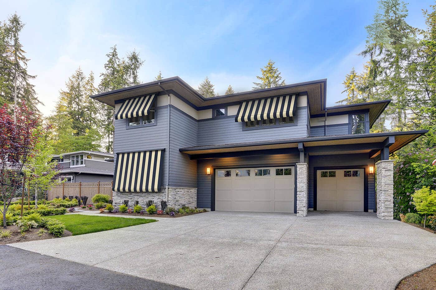 Outside view of a blue mid-century style home with a garage, surrounded by trees, featuring Luxaflex Re-covering Awnings. Available in various styles and durable fabrics.