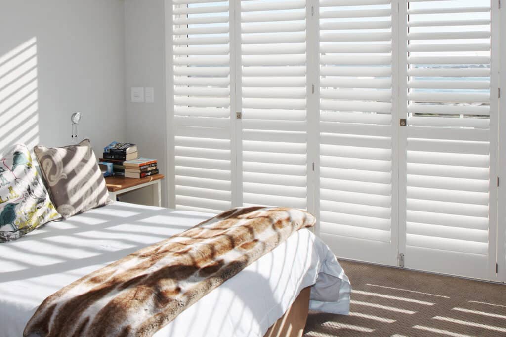 Cozy and cool white bedroom featuring Norman Brightwood Shutters. Sunlight streaking through the shutters. Shutters for Sliding Doors.